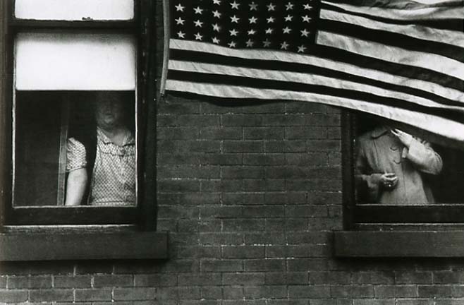 Two figures at windows above a brick wall, one face in shadow, the other hidden behind a large American flag hanging across the frame.