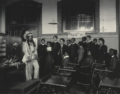 A classroom of students in ordered rows, a figure in Native American dress standing at the front beside a display of artefacts.