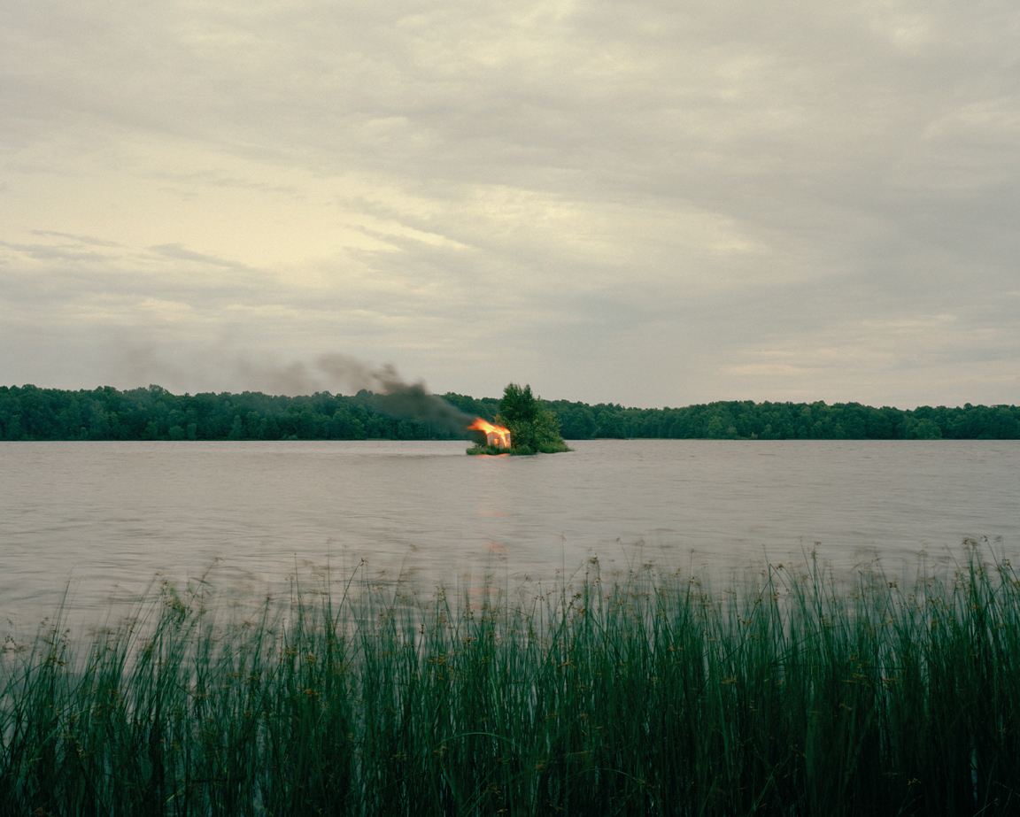 A small wooden house alight on a tiny island in a lake, reflected in the water.